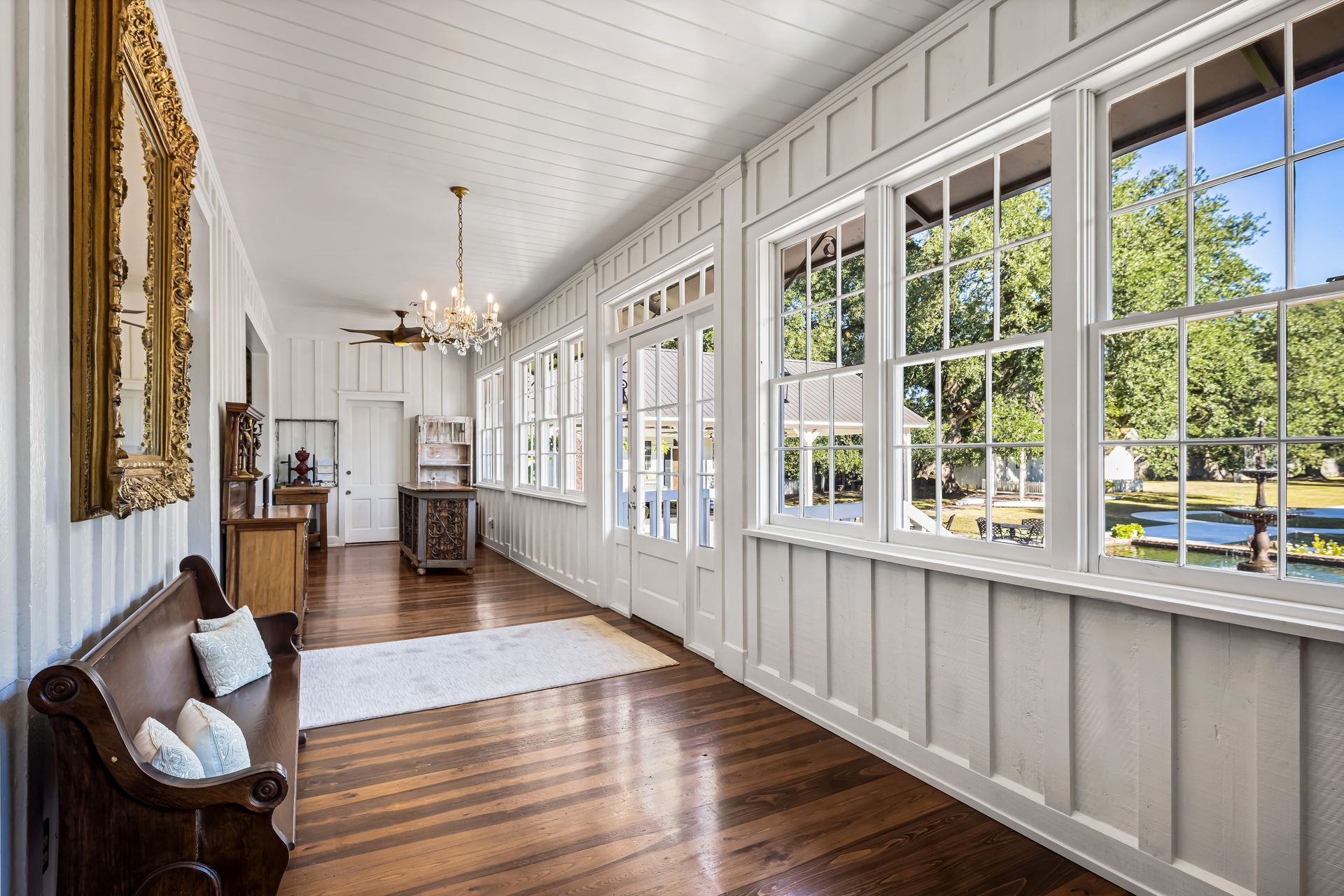 Long hallway with white paneled walls, wood floors, windows overlooking trees, and antique furniture.