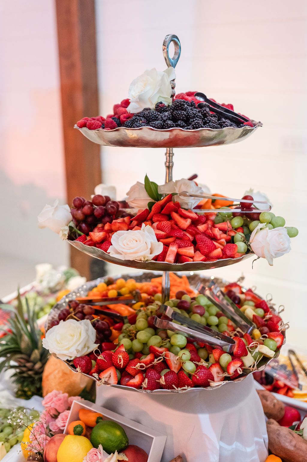 Three-tiered fruit platter with berries, grapes, and strawberries, decorated with flowers on a white table.