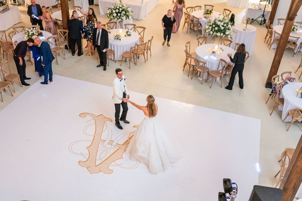 Bride and groom on a dance floor with a monogram. Guests watch from tables in a decorated venue.