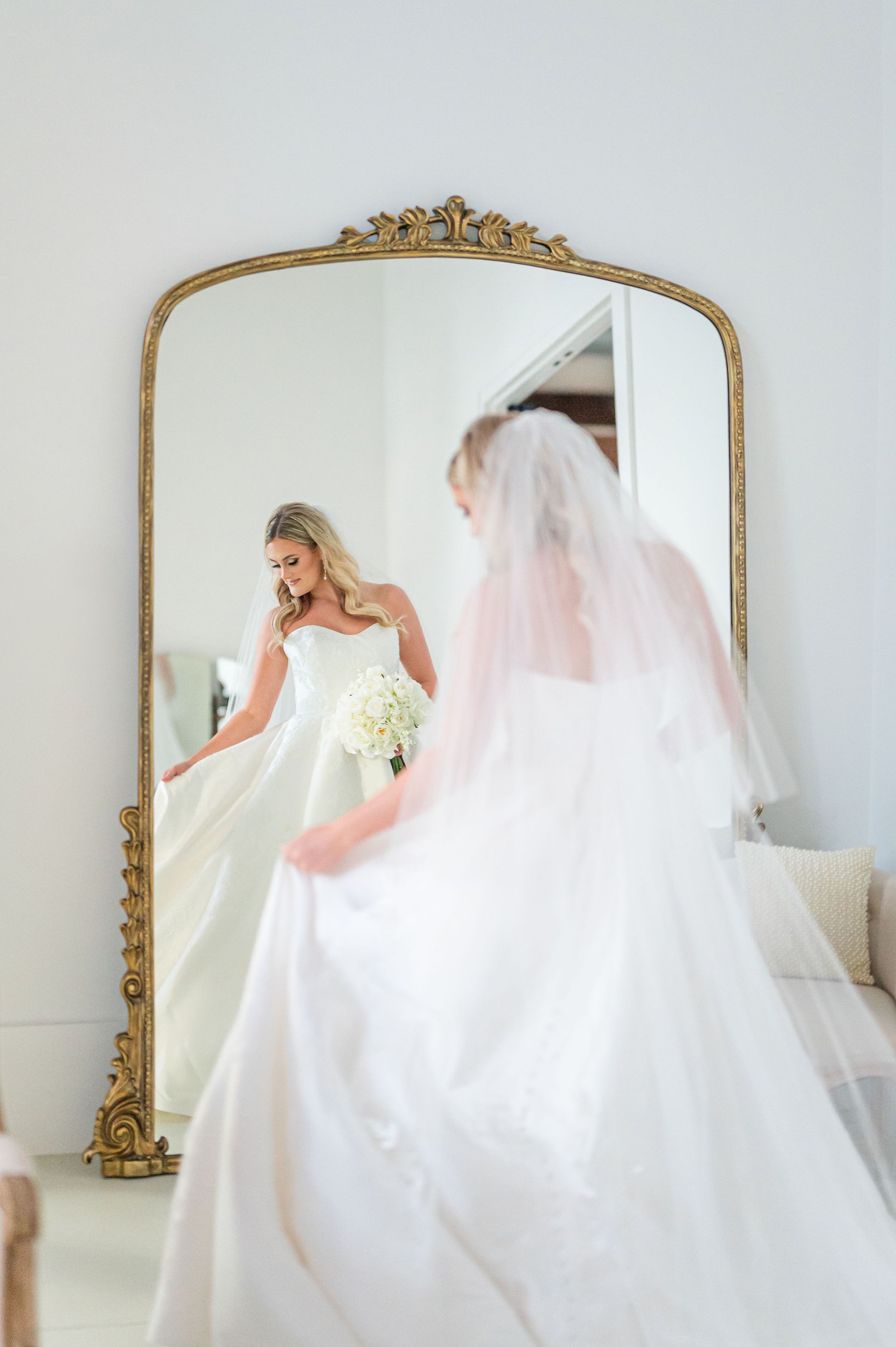 Bride in wedding dress, looking in ornate gold-framed mirror, holding bouquet.