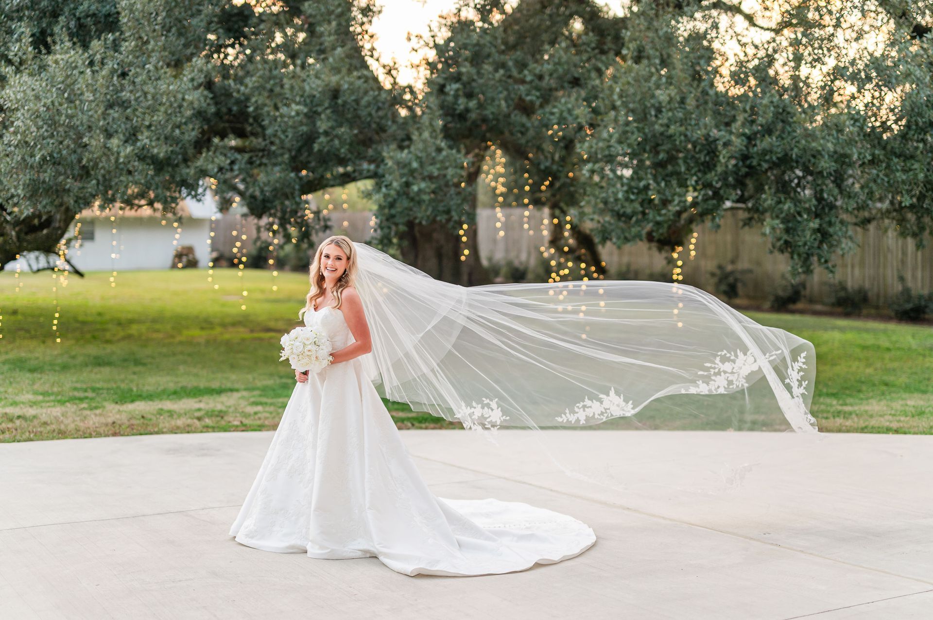 Bride in white gown, veil blowing in the wind, holding flowers, standing on patio, trees and lights in background.
