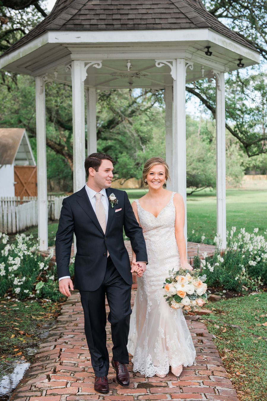 Bride and groom walk hand-in-hand on brick path under a gazebo; groom in suit, bride in lace dress, holding bouquet.