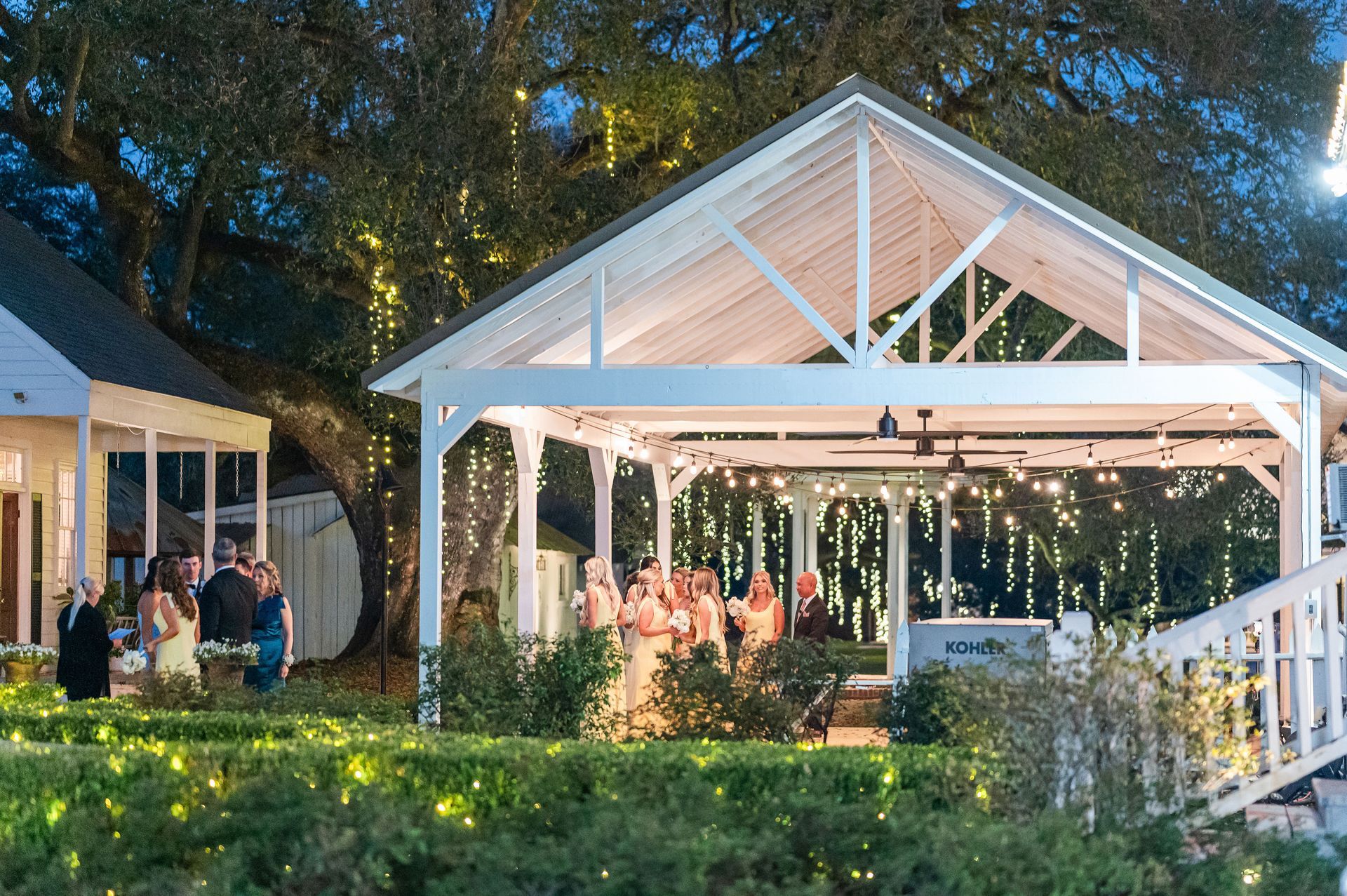 Wedding ceremony under a white gazebo, with guests standing on a lawn at dusk, string lights are lit.