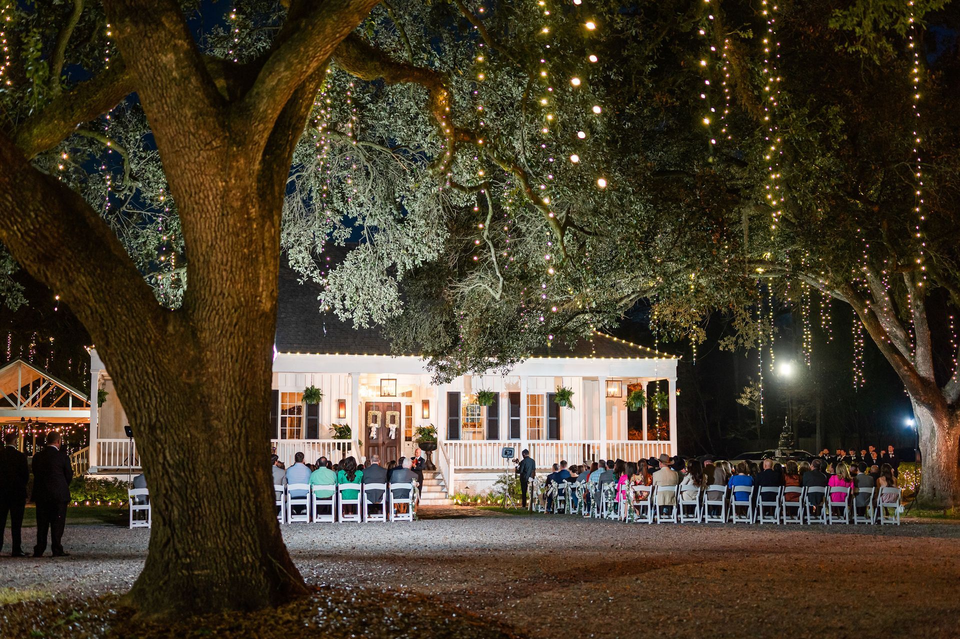 Nighttime outdoor wedding ceremony under large trees strung with lights, white house in the background.