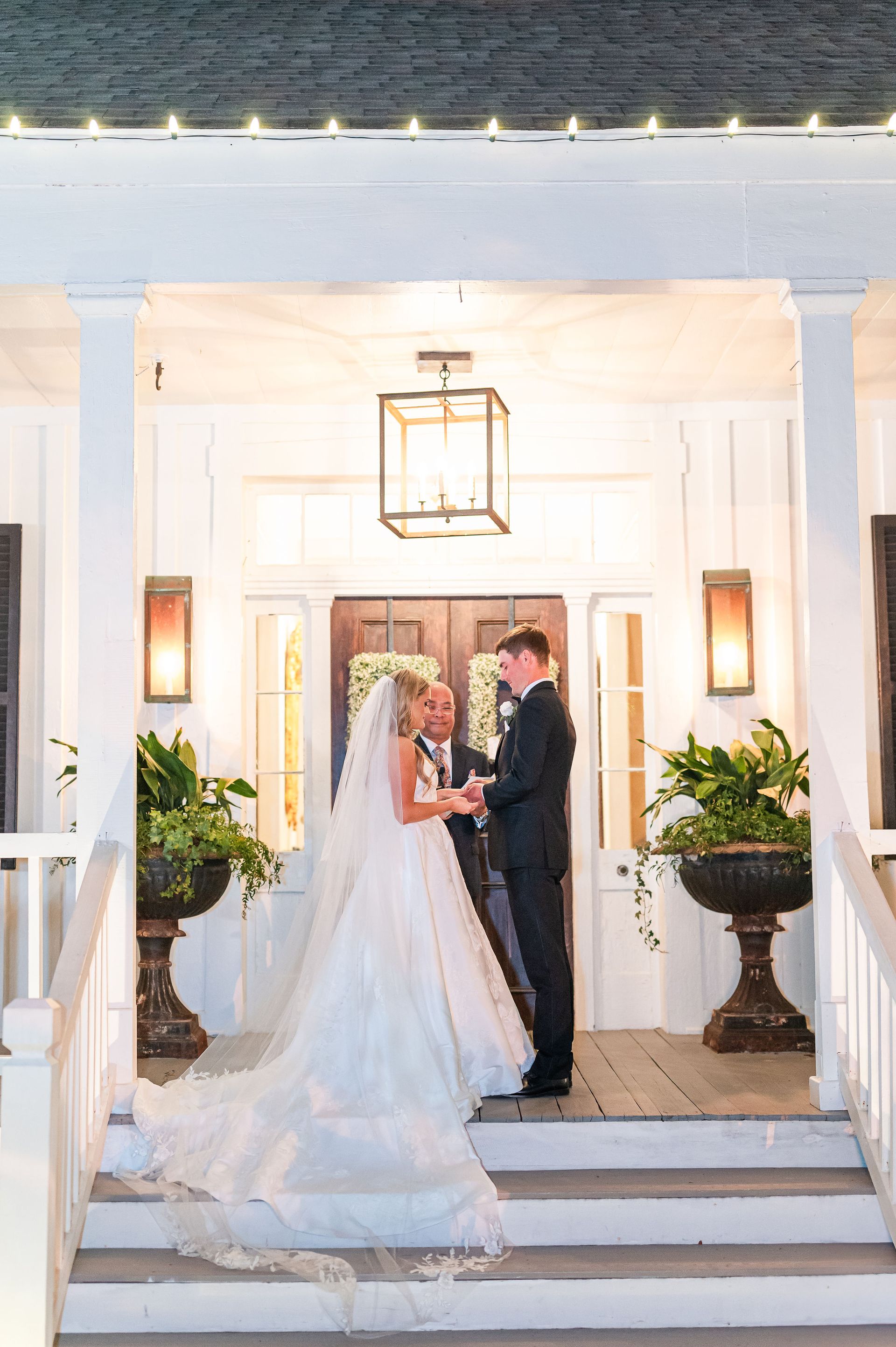 Couple at wedding ceremony, bride in white gown, groom in suit on porch with flower pots.