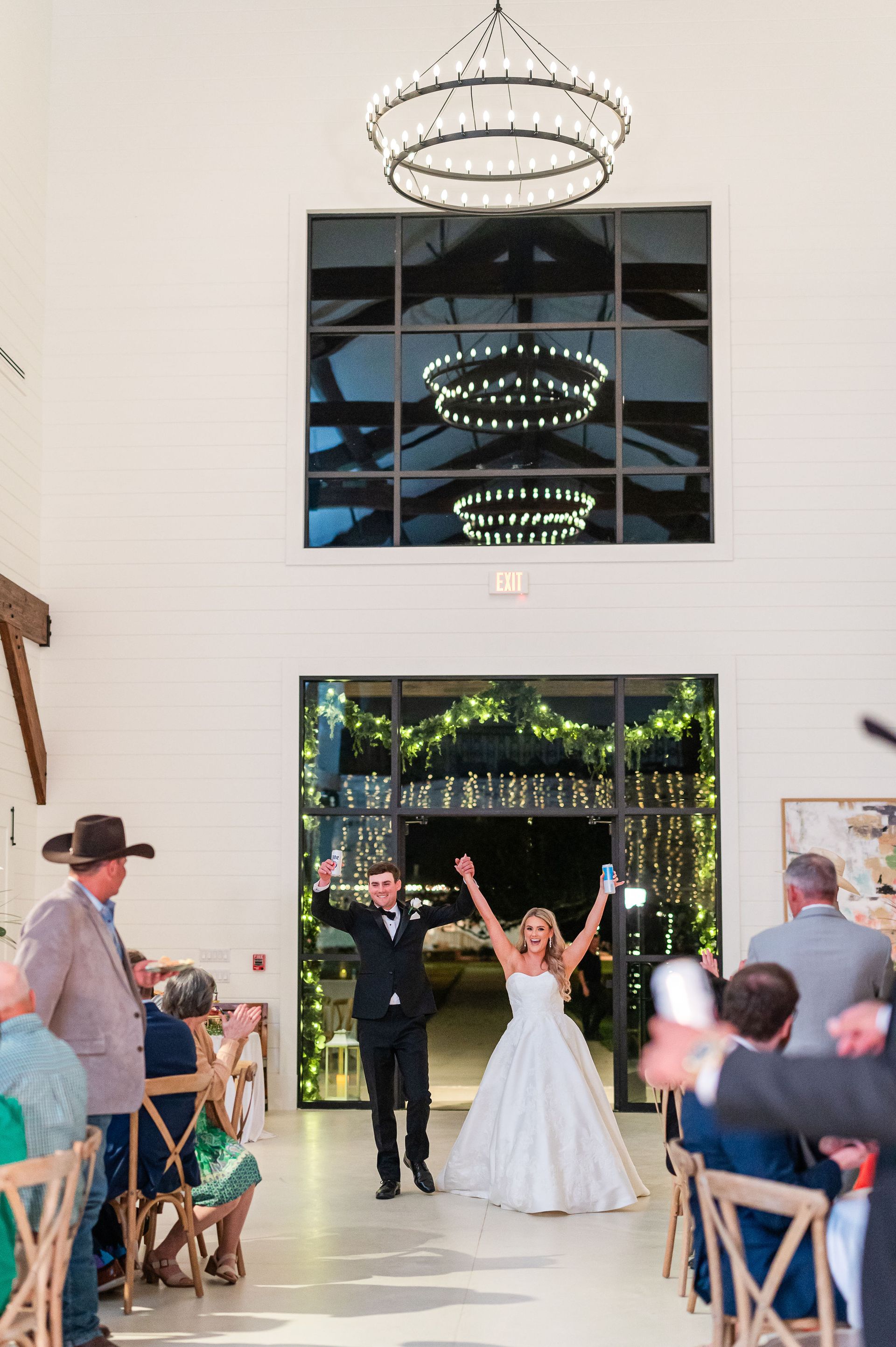 Bride and groom exiting wedding, arms raised in joy. Indoor ceremony space with large window, chandelier.