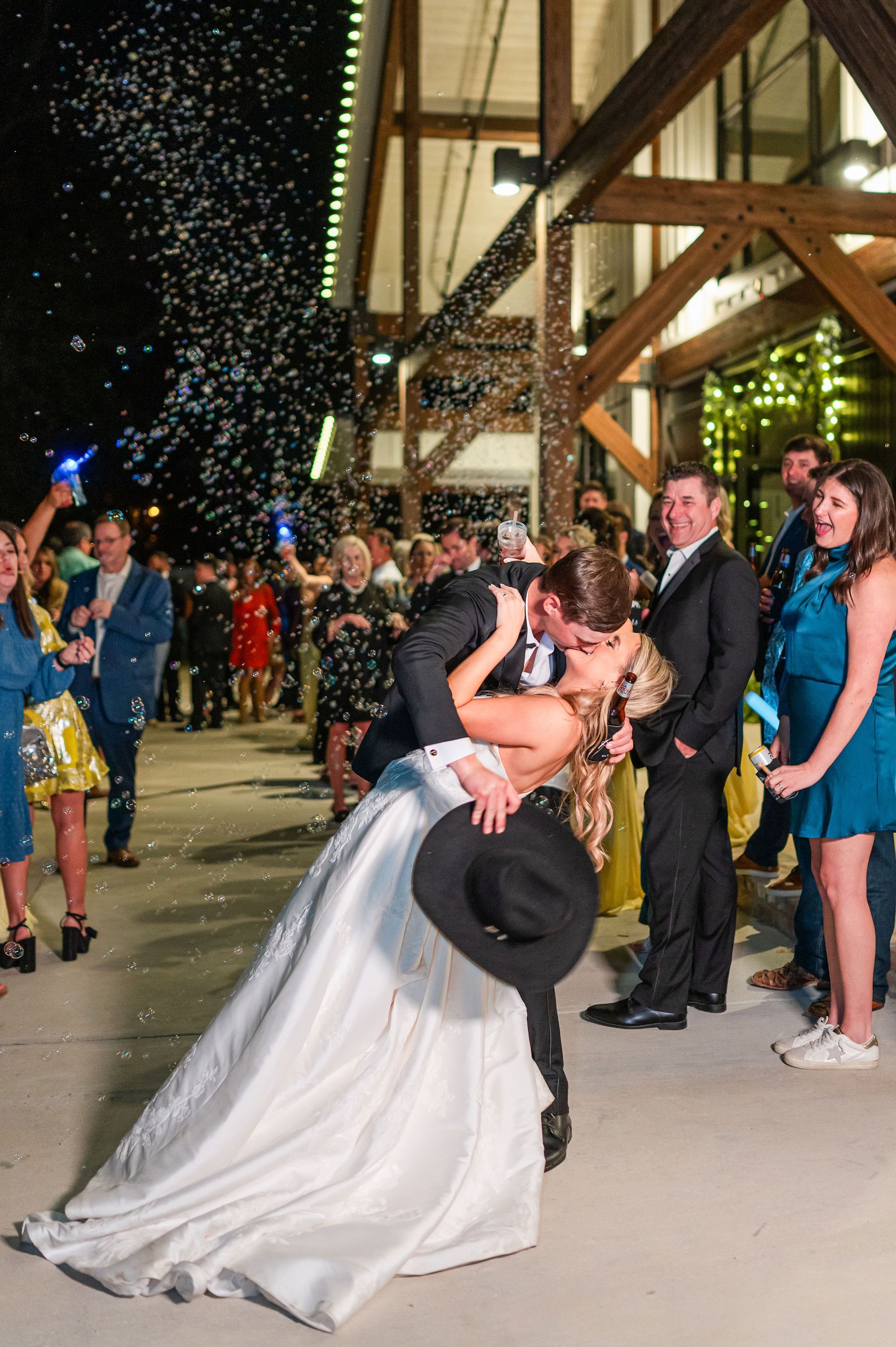 Newlyweds kiss, confetti falls. Bride in white gown, groom in black suit, guests cheer outside a building at night.