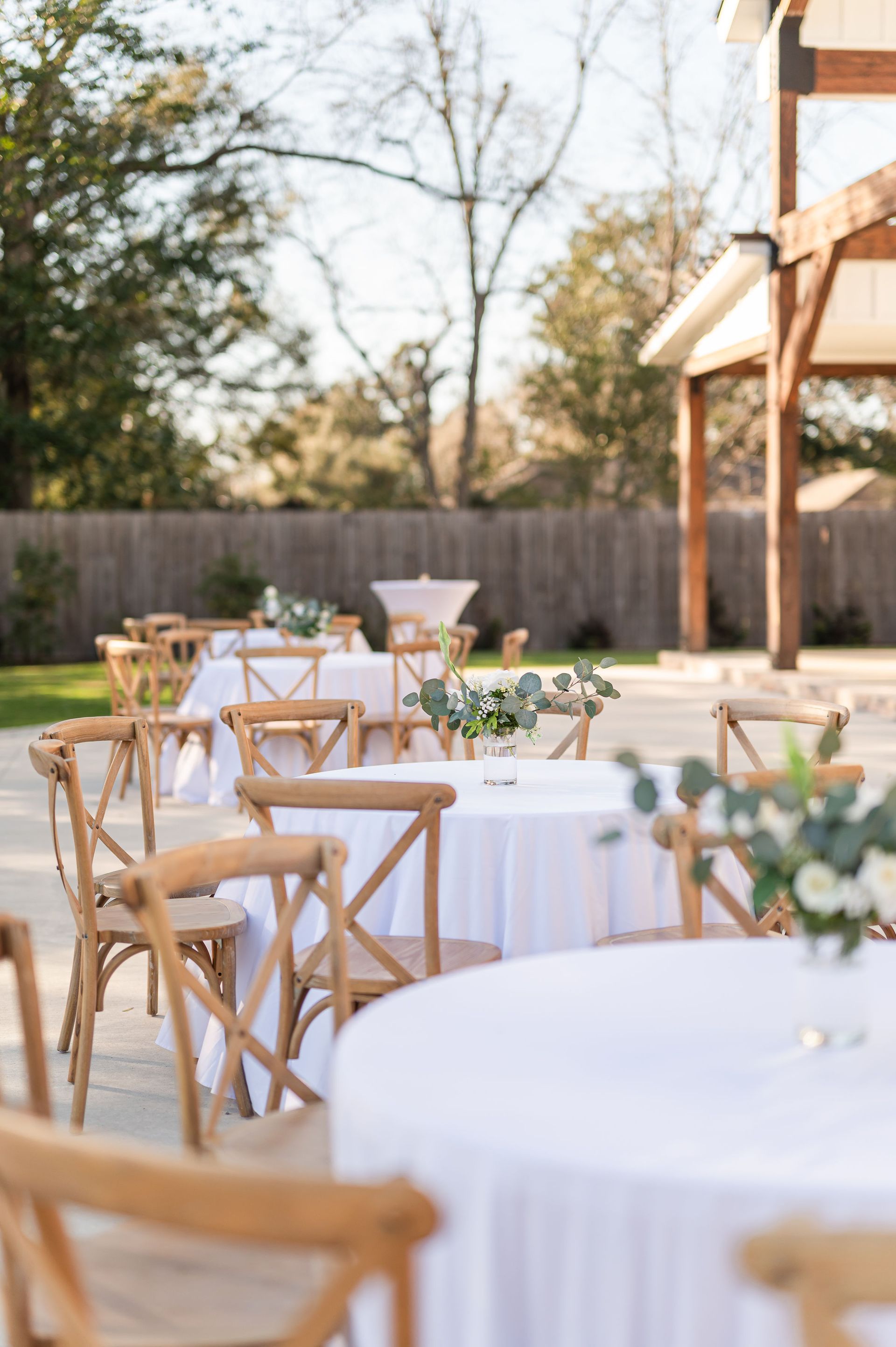 Outdoor event tables with white tablecloths and floral centerpieces; cross-back chairs.