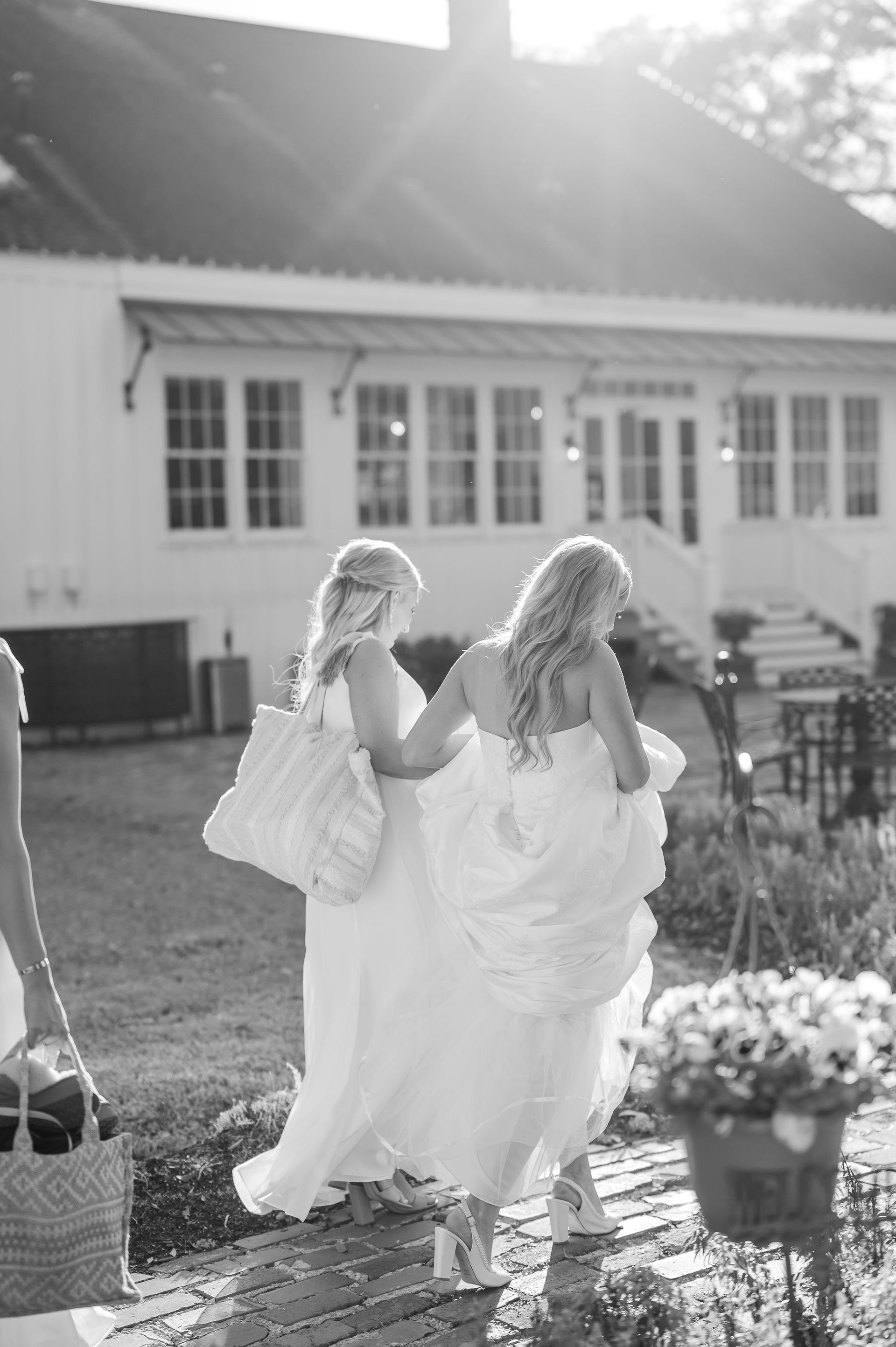 Two women in white dresses walk away, holding hands, near a white building in sunlight.