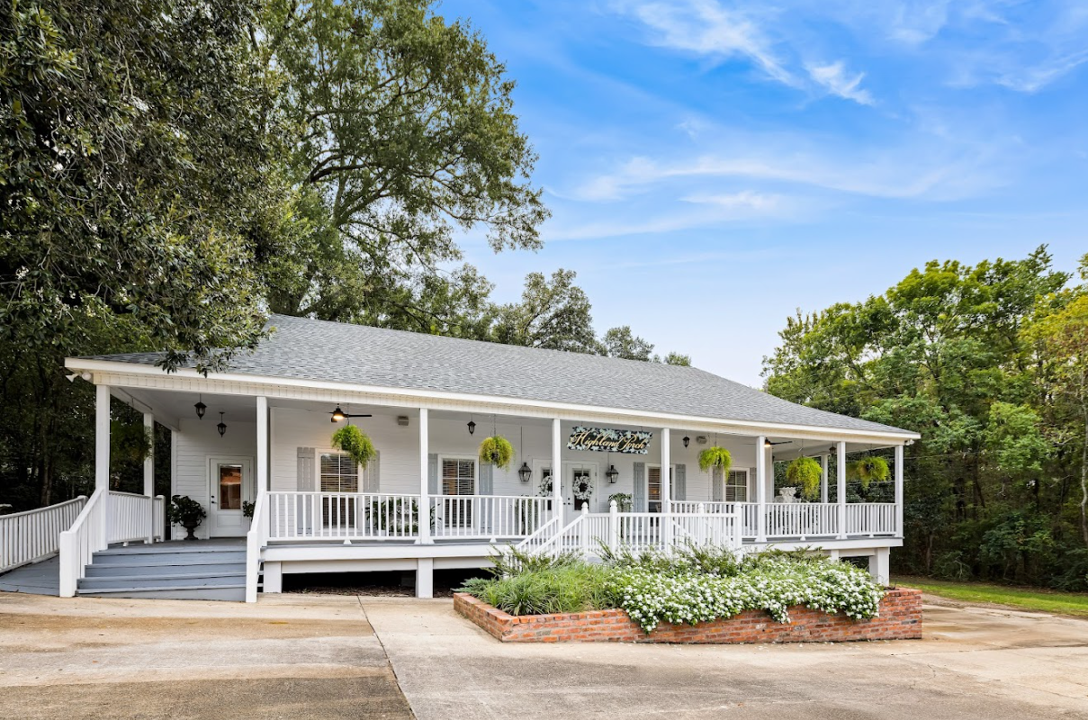 White house with wraparound porch and gray roof, surrounded by trees and landscaping.