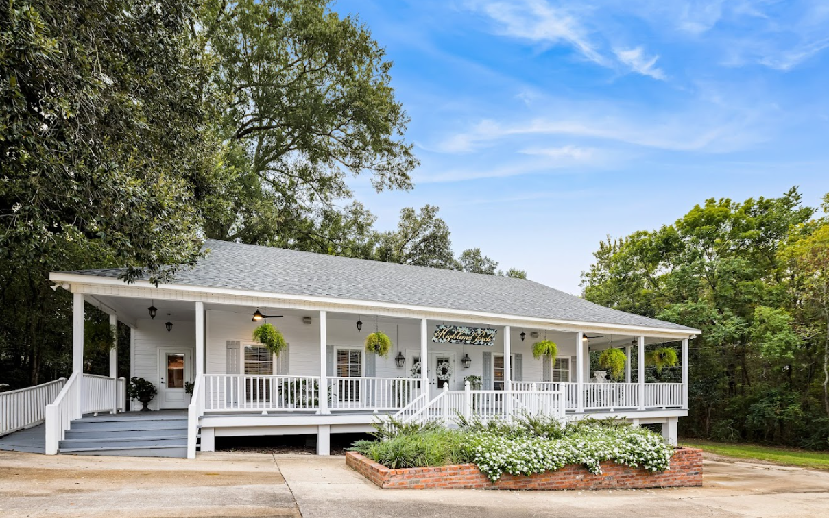 White house with porch and flowerbeds, blue sky, and trees.