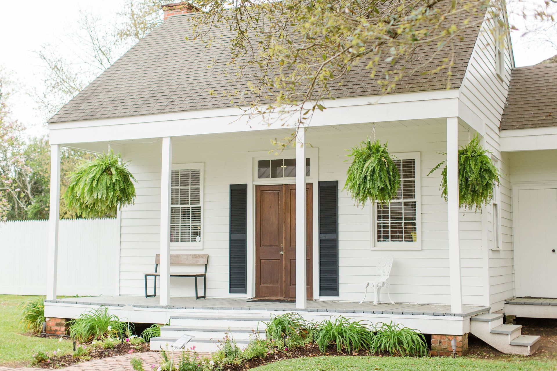 White house with porch, brown door, black shutters, green hanging ferns, stone steps, and grass.