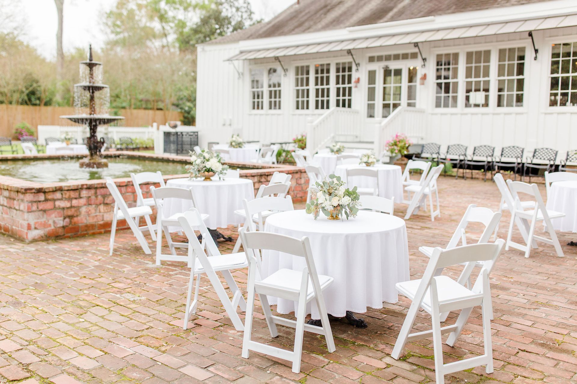Outdoor event with round tables, white chairs, and floral centerpieces. A brick patio with a fountain and a white building.