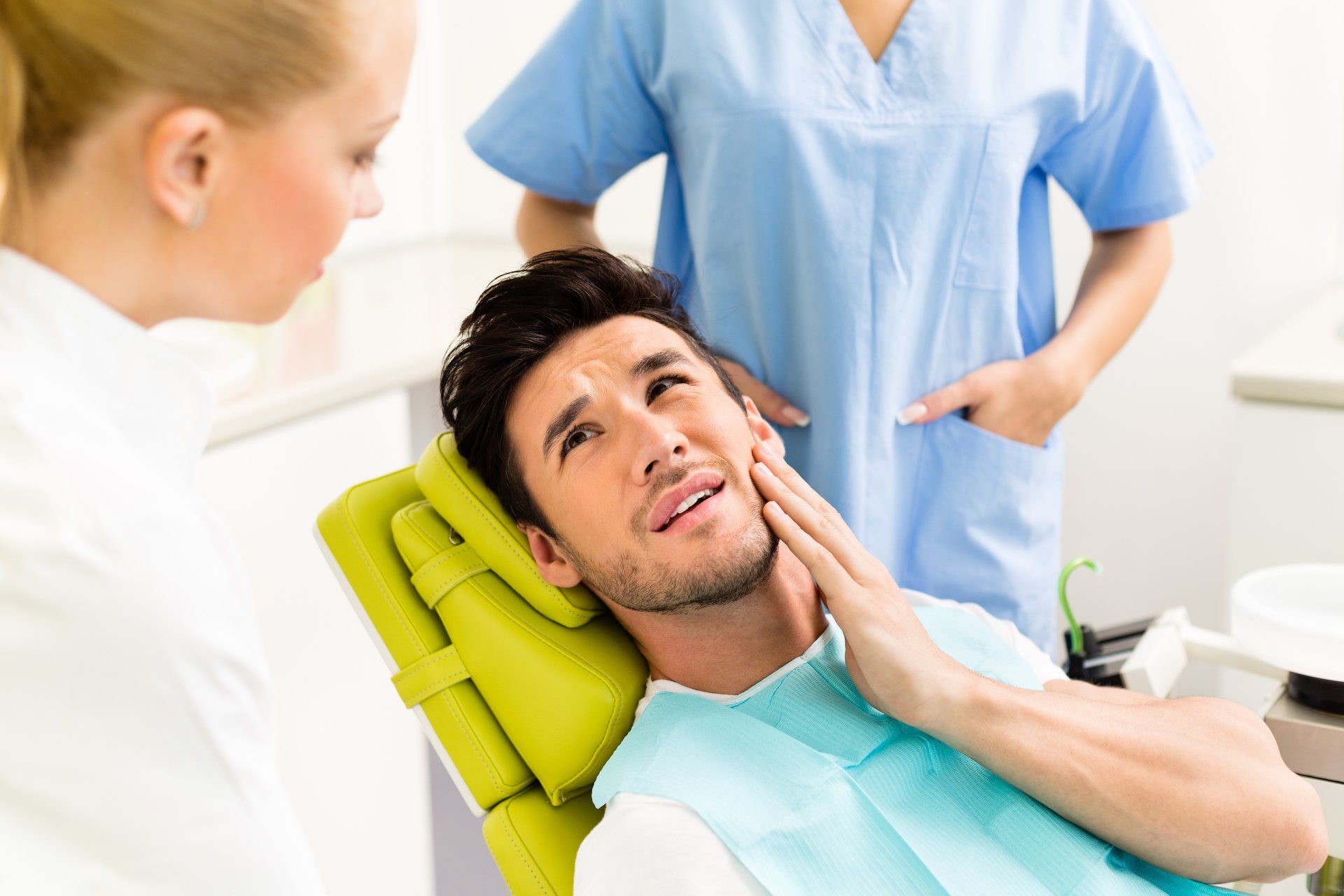 Dental patient in green bib holding his cheek while dentist and assistant stand nearby in a clinic