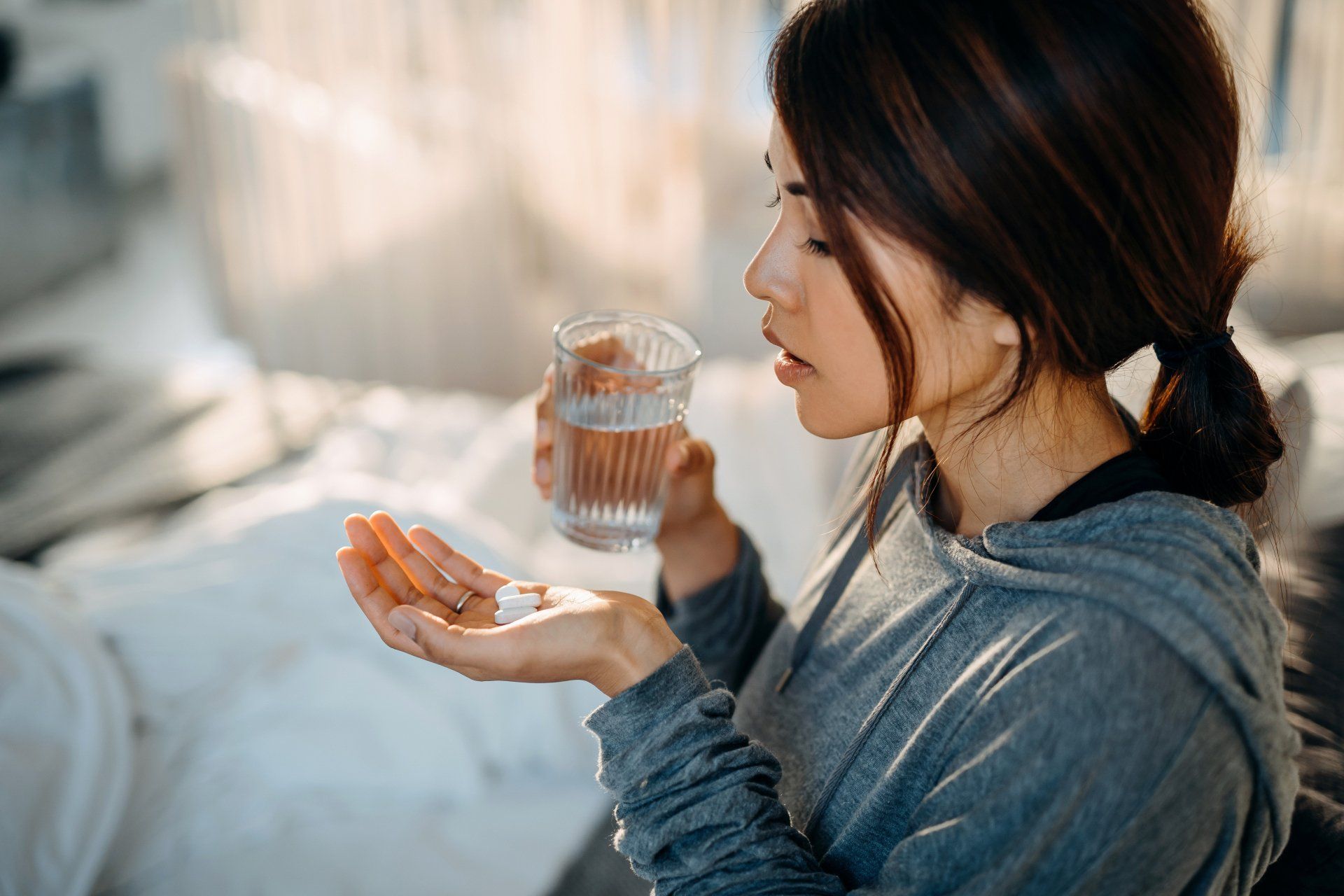 Woman holding a glass of water and a pill beside a bed, looking at her hand