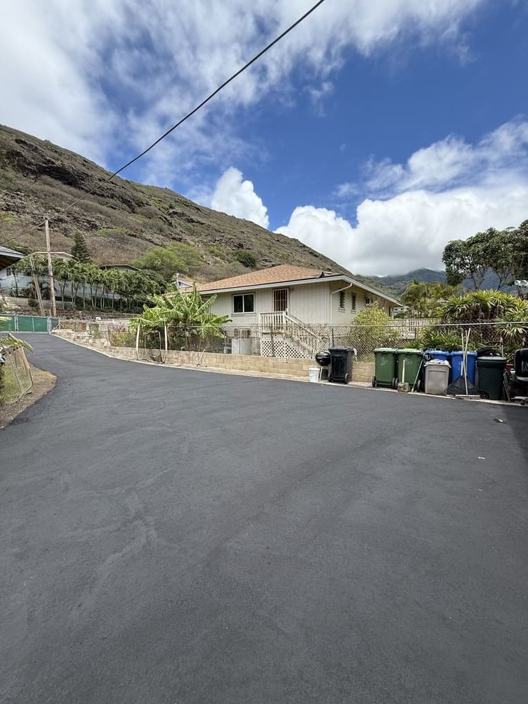 House with asphalt driveway, trash cans, and mountain backdrop under a partly cloudy sky.