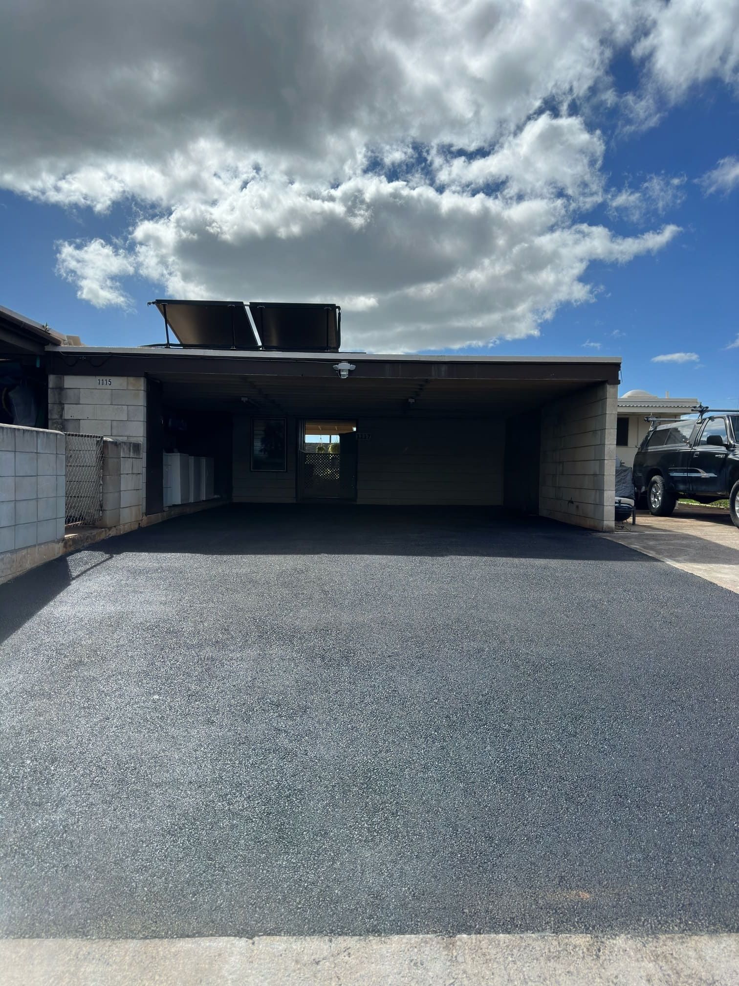 Asphalt driveway leading to a carport with a solar panel on the roof, under a cloudy blue sky.