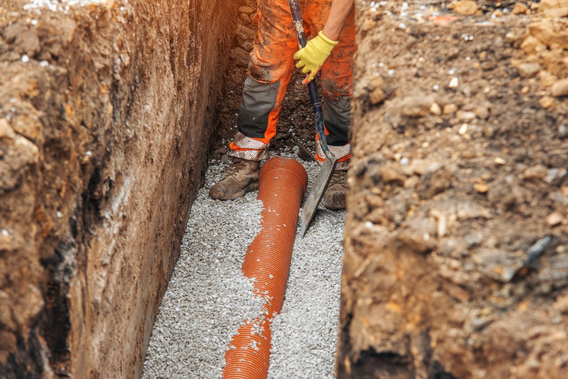 Worker installing orange corrugated pipe in a trench with gravel bed and dirt walls