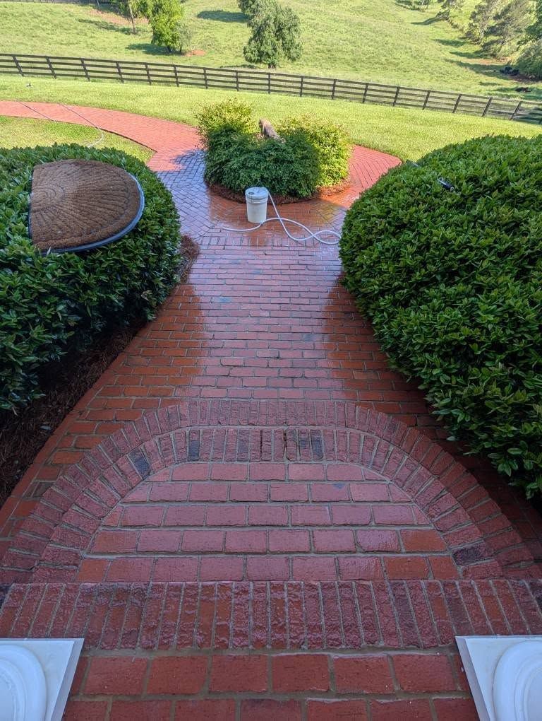 Brick pathway and stairs flanked by green bushes leading toward a grassy field with a wooden fence.