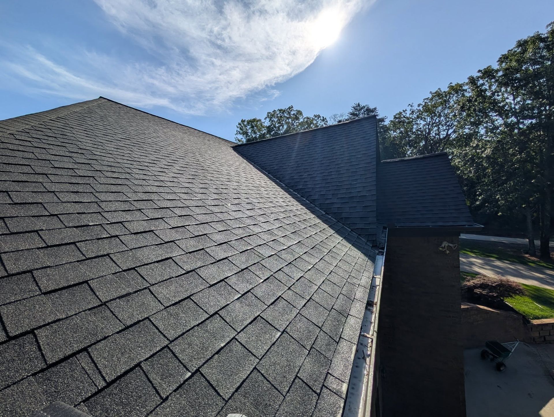 Dark gray shingled roof with a gutter, under a partly cloudy blue sky. Trees and sunlight are visible.