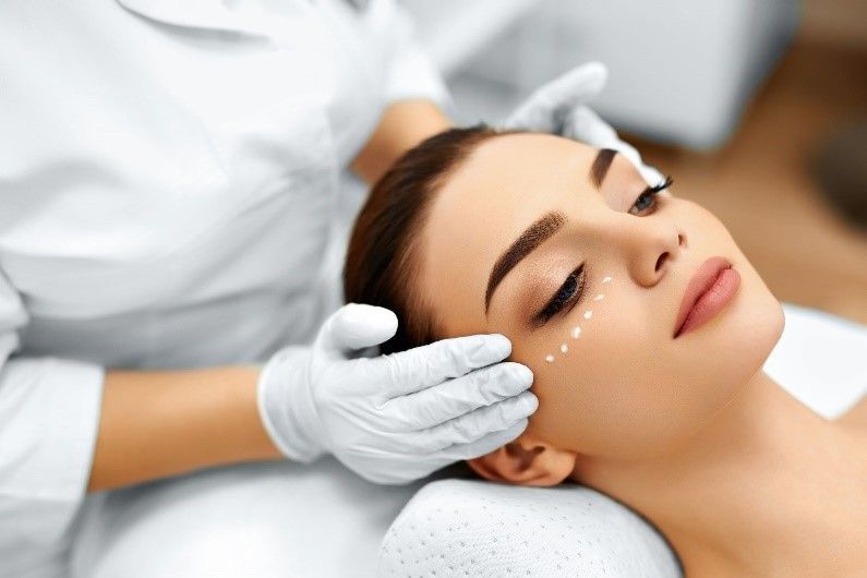 Woman receiving cosmetic procedure, white gloves hold her face; dotted lines mark treatment area.