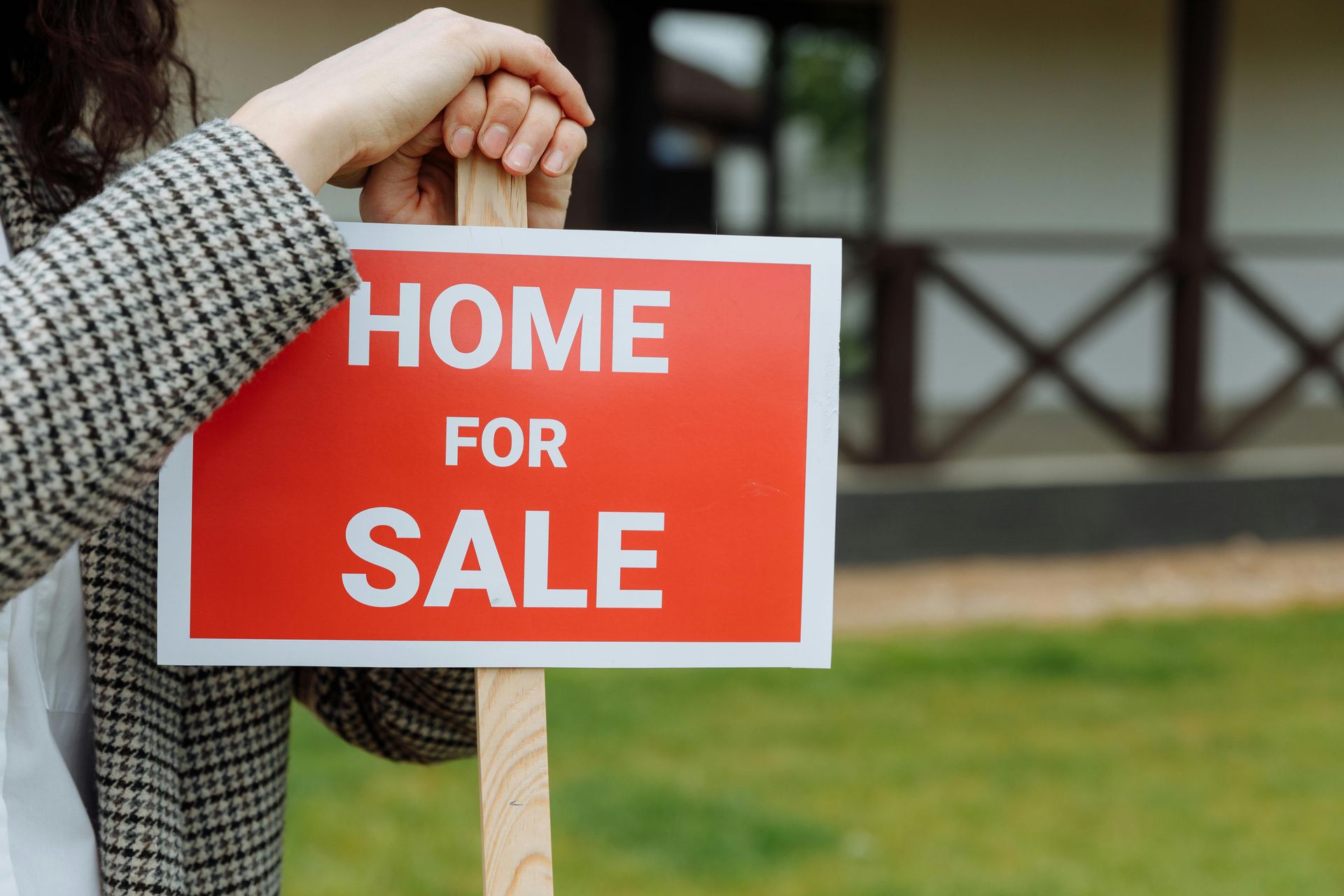 Hand holding a red “HOME FOR SALE” sign in front of a house and lawn