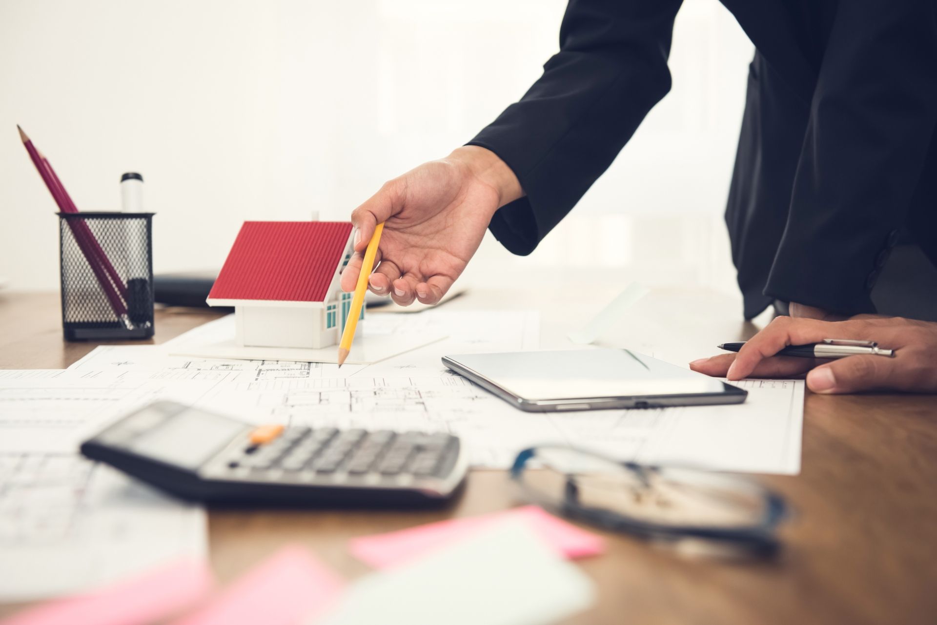 Hand pointing a pencil at documents on a desk with calculator, clipboard, and model house in an office setting