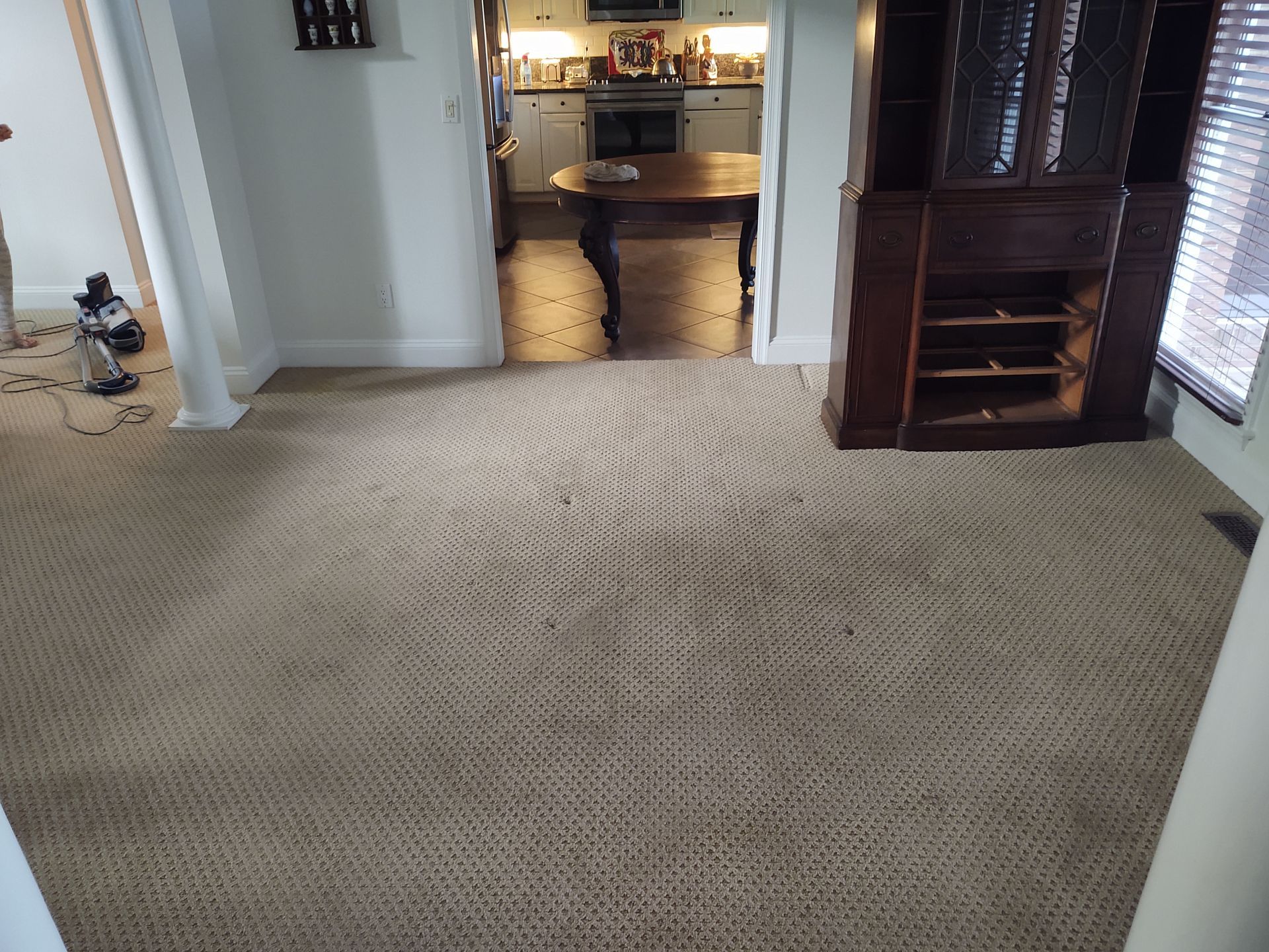 A carpeted room with a dark wooden hutch, leading to a kitchen with a dining table and light-colored cabinets.
