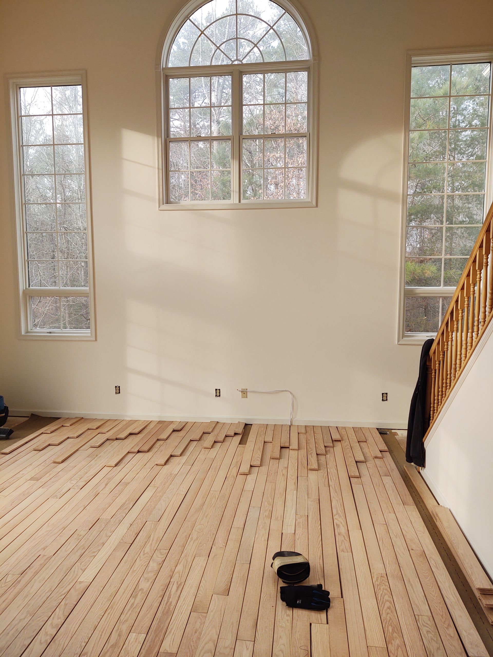 A room under renovation with new, light-colored unfinished wood floor planks being laid down in front of three windows.