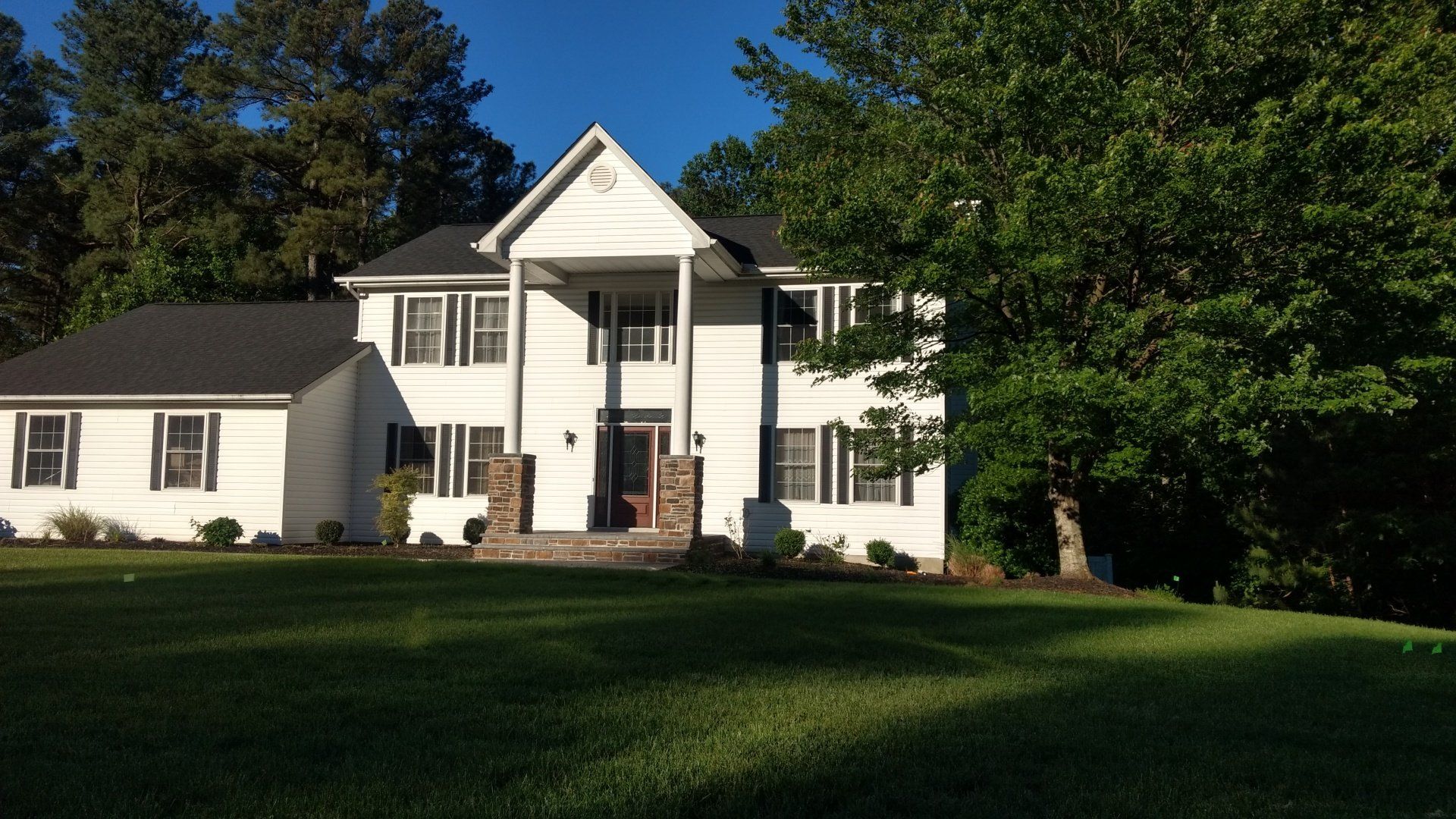A two-story white house with dark shutters and a central covered porch, set on a green lawn surrounded by tall trees.