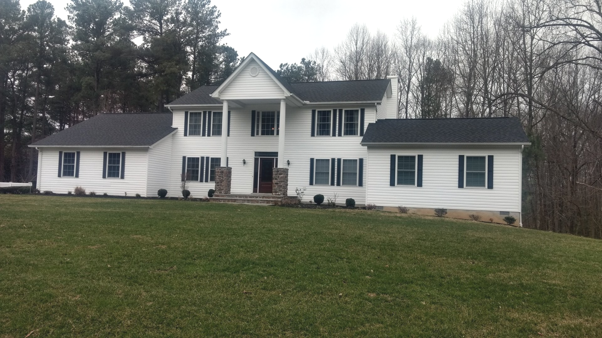 Large white two-story house with black shutters on a grassy hill under an overcast sky