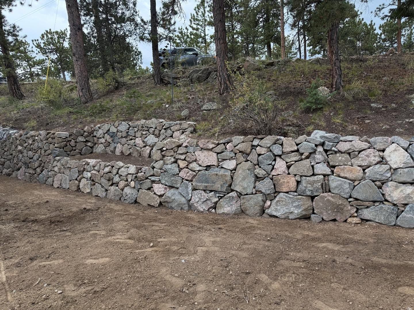 Stone retaining walls built into a hillside, trees in the background, dirt foreground.