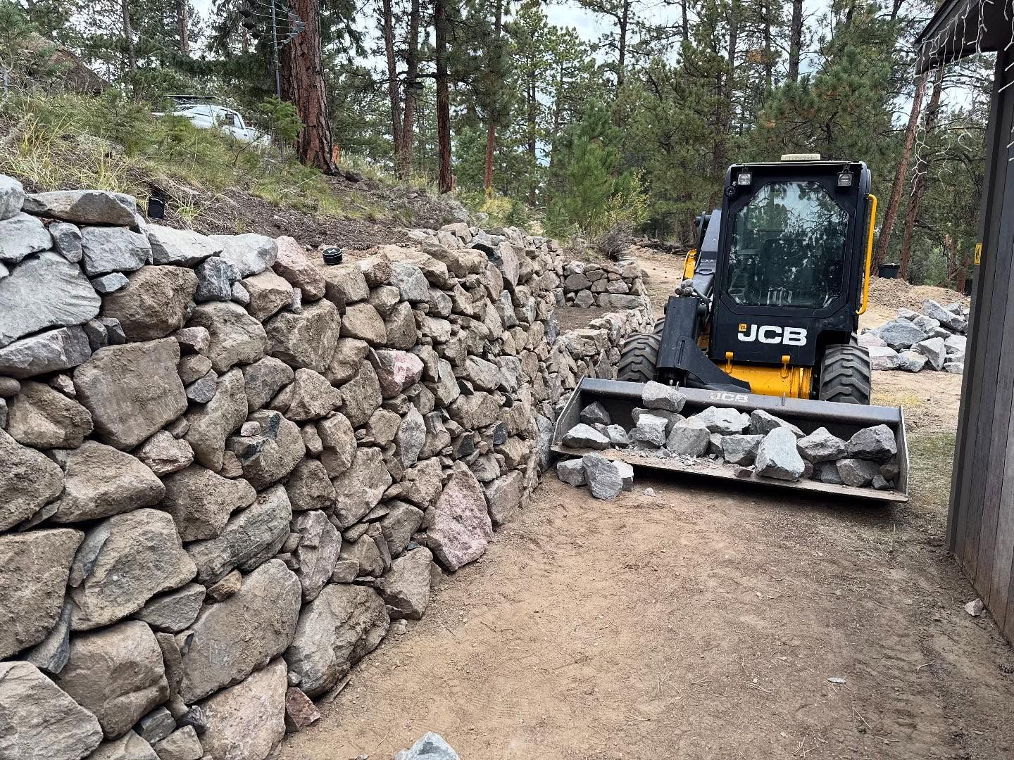 Bobcat loader moving rocks next to a stone wall; construction in a wooded area.