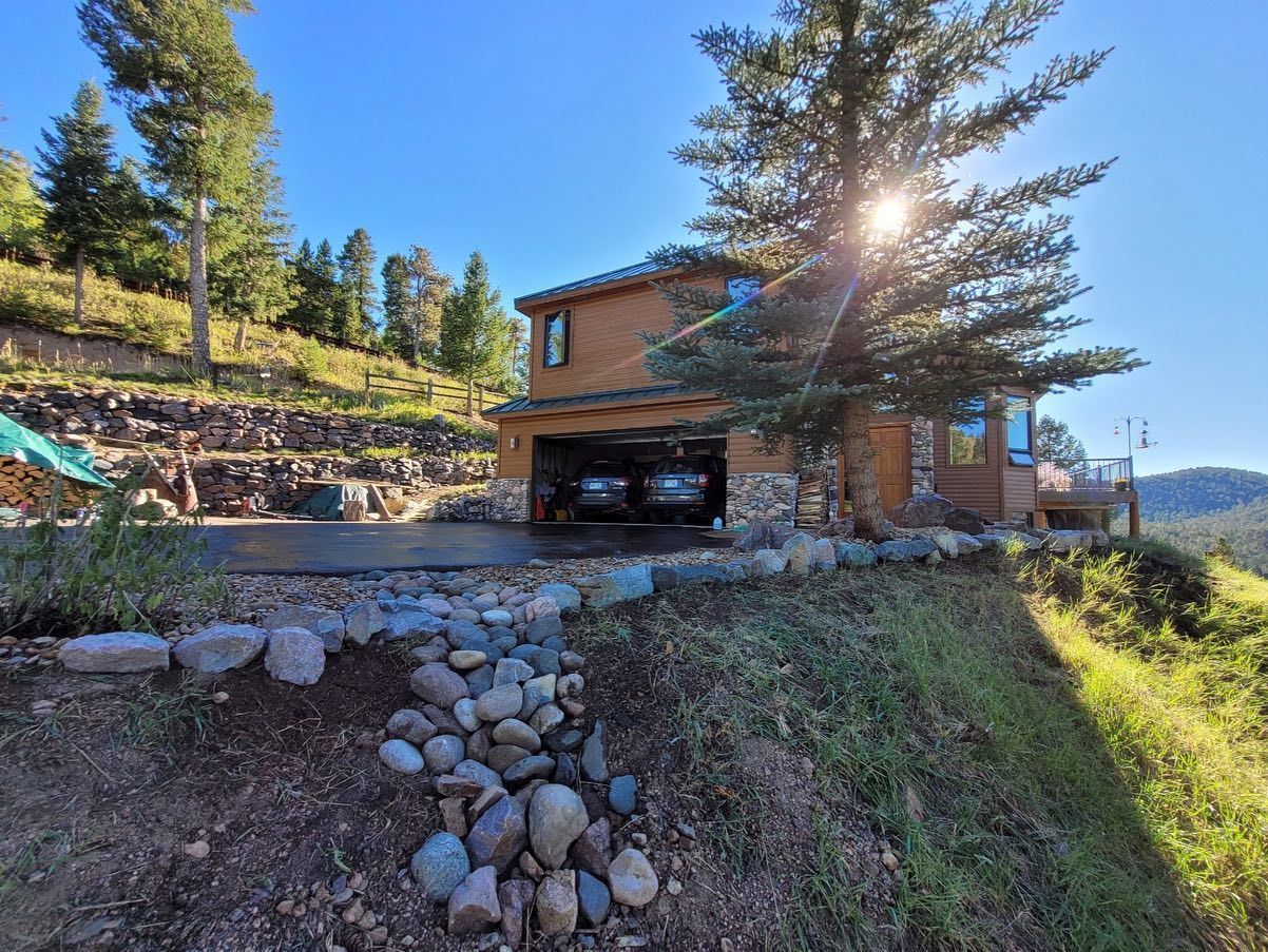 House with a garage and two cars, sunlight shining through a tree, rocks in foreground.