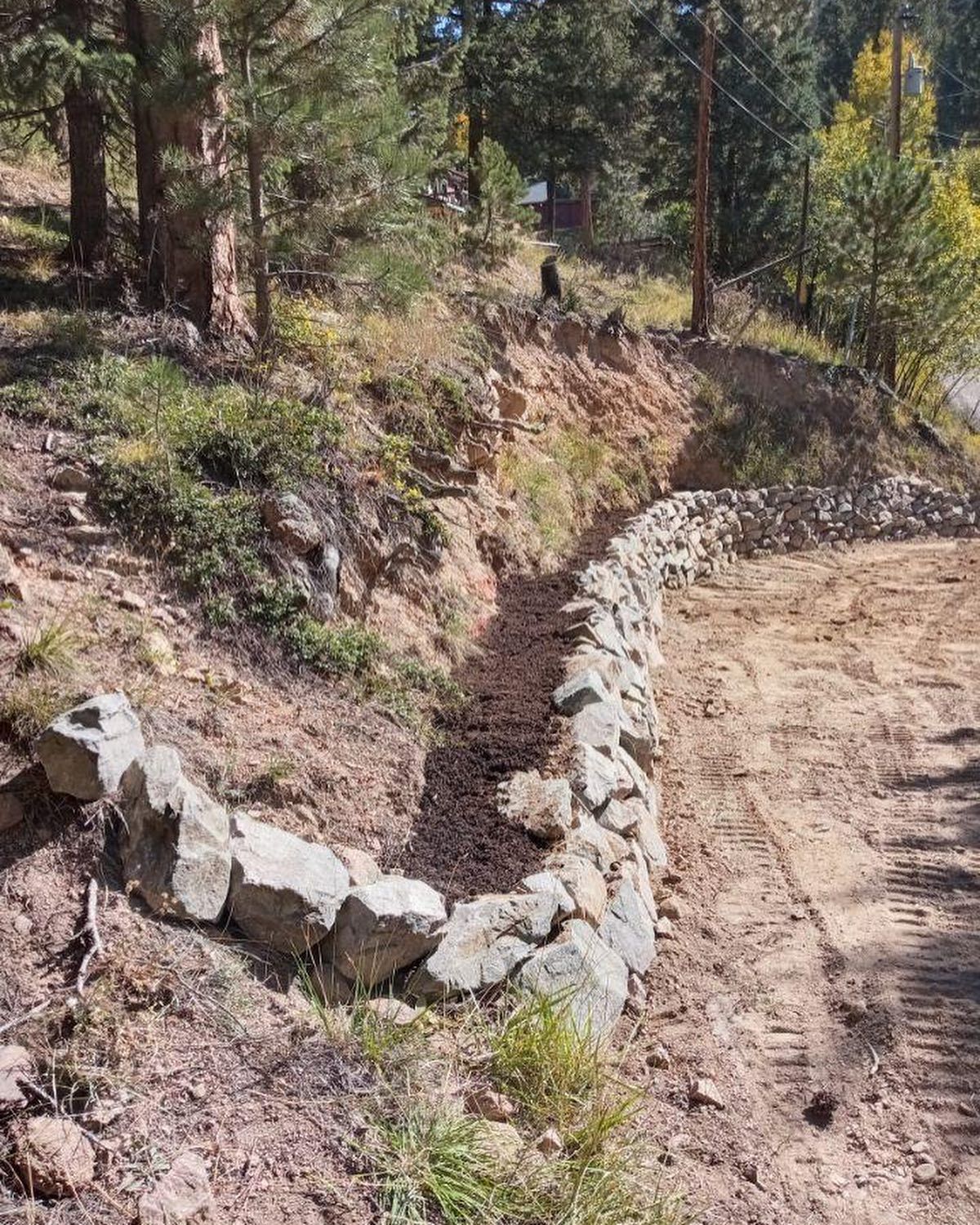 A rock retaining wall curves along a dirt road, likely for erosion control, in a wooded area.
