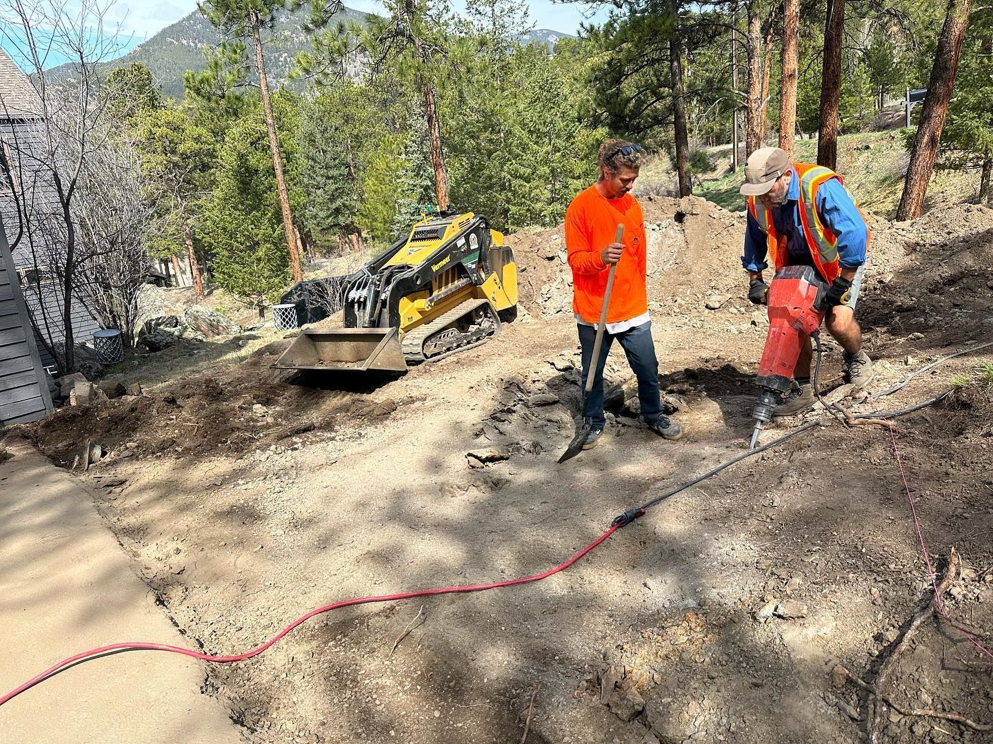 Two workers using equipment on a dirt path in a wooded area. One uses a jackhammer. A small excavator sits nearby.