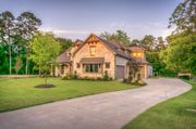 Stone house with a long driveway, green lawn, and trees under a blue sky.