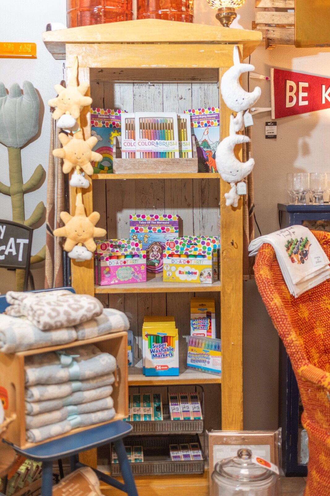 Wooden shelf displays art supplies and soft toys in a shop, with a blue crate of blankets below.