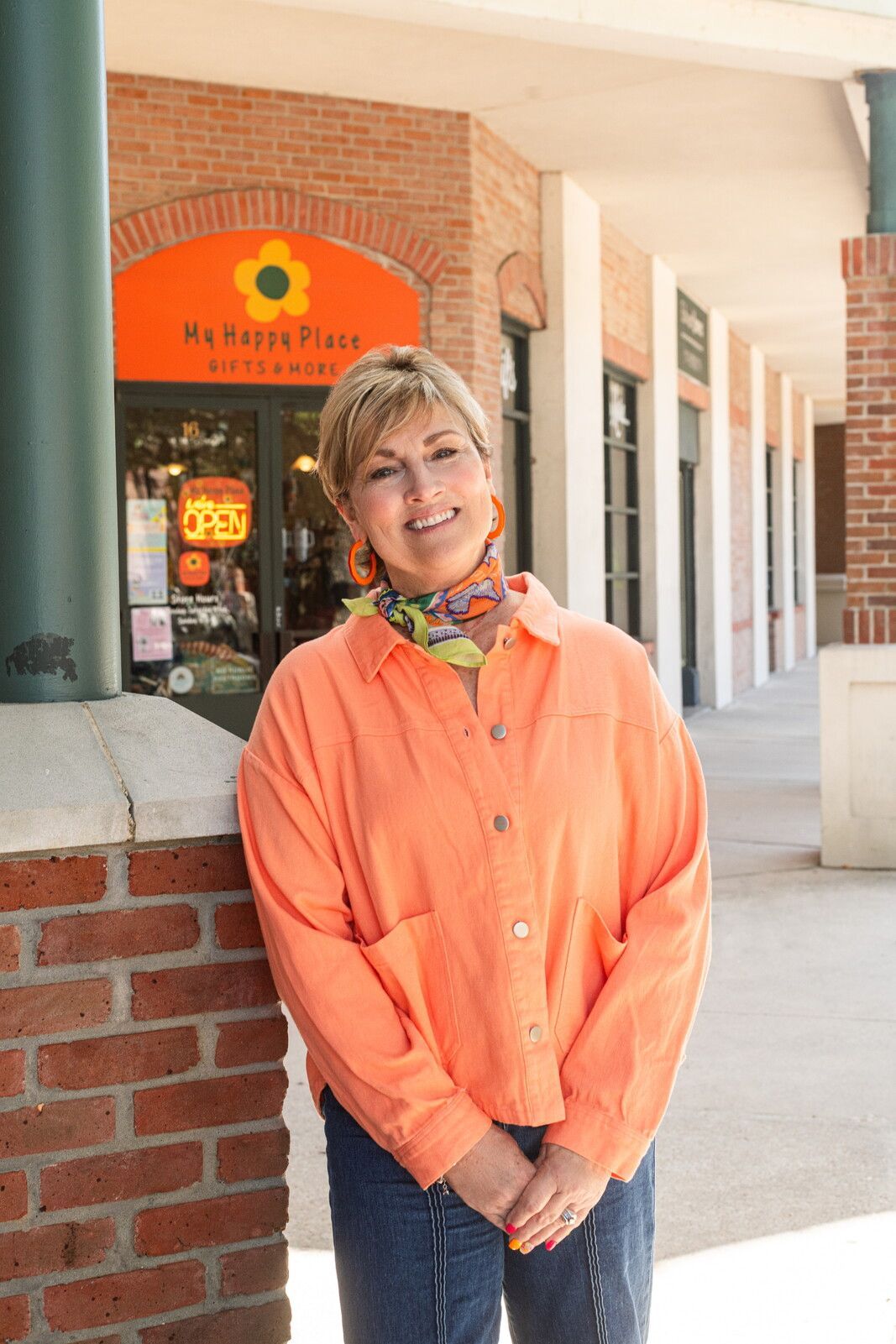 Woman in orange shirt smiles in front of a store, wearing colorful earrings.