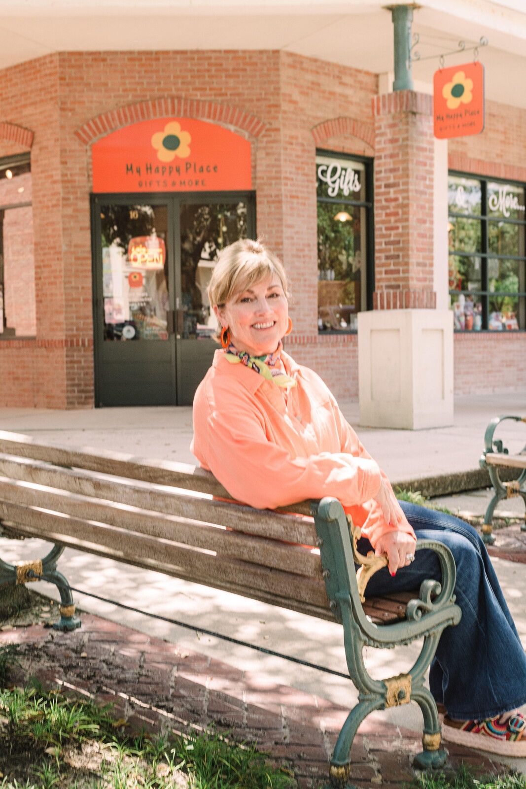Woman sitting on bench in front of a store with a sunflower sign; orange shirt, jeans, smiling.