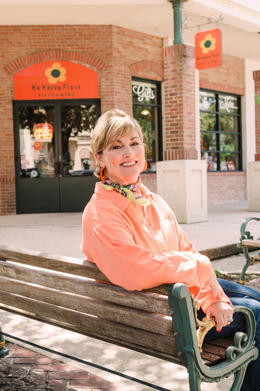 Woman in orange shirt sits on a bench outside a shop with a sunflower logo.