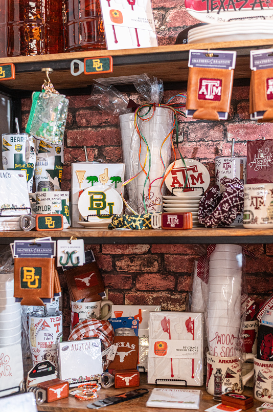 Shelves displaying college-themed merchandise, including mugs, coasters, and napkins, against a brick wall.