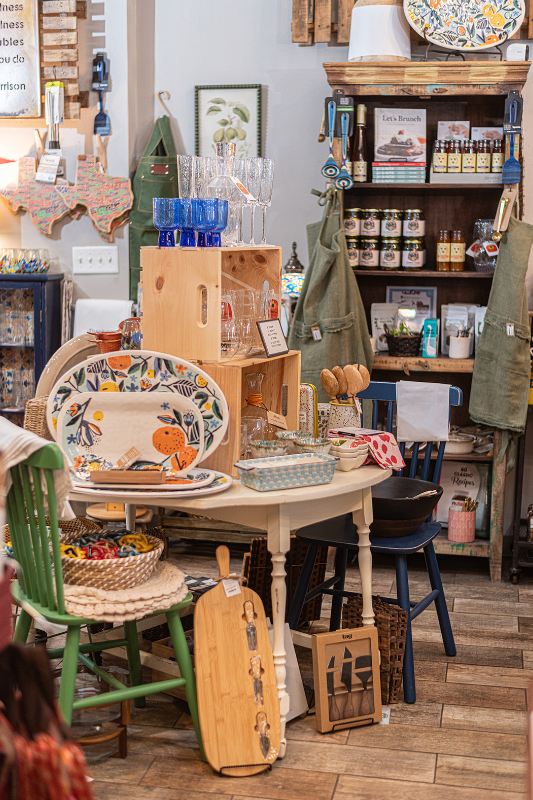 Interior shop display: wooden shelves with jars, glassware, and serving dishes on a round table and stools.