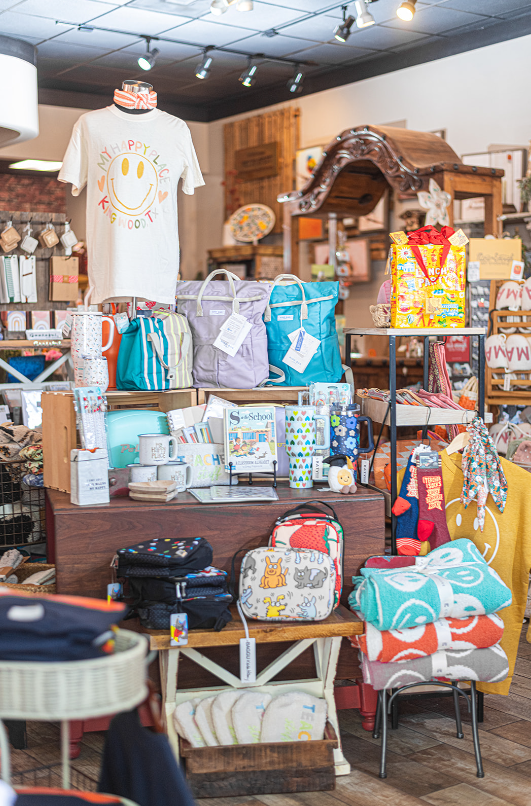 Inside a gift shop, display of bags, mugs, shirts, and home goods. Bright colors, various patterns, wooden furniture.