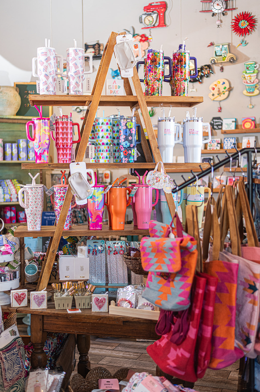 Wooden display stand with mugs and tumblers in a shop. Colorful bags and accessories hang on a rack.