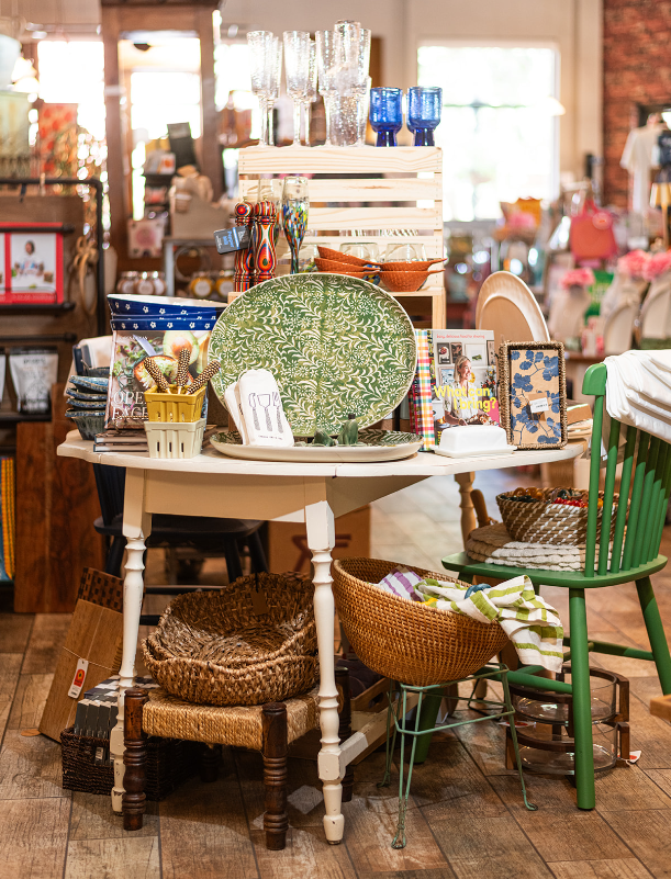 A retail display of dishes, glassware, and decorative items on a white round table, in a store with wood floors.
