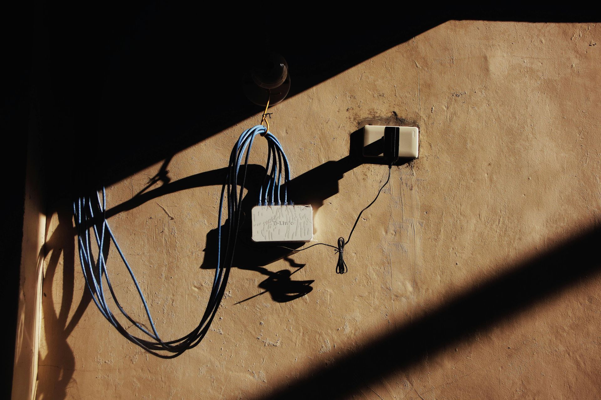 Coiled blue cable and rectangular block on a textured tan wall, illuminated by strong sunlight casting dark shadows.