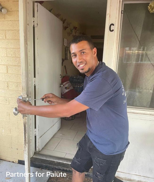 Man opening door, smiling. He wears a blue shirt and black shorts. Standing outside a building with the letter 