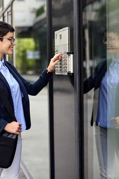 Woman in a blue blazer entering a code on a wall-mounted keypad beside a glass door