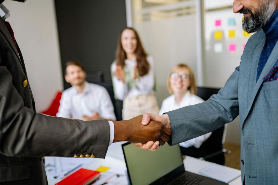 Two people shaking hands in a bright office meeting room, with colleagues seated in the background.